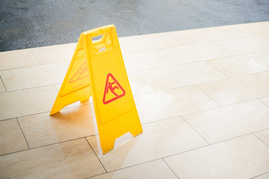 A bright yellow wet floor caution sign featuring a red triangle with a person slipping symbol, positioned on a beige tiled floor in an indoor setting. The sign is placed near the edge of the tiles, wh