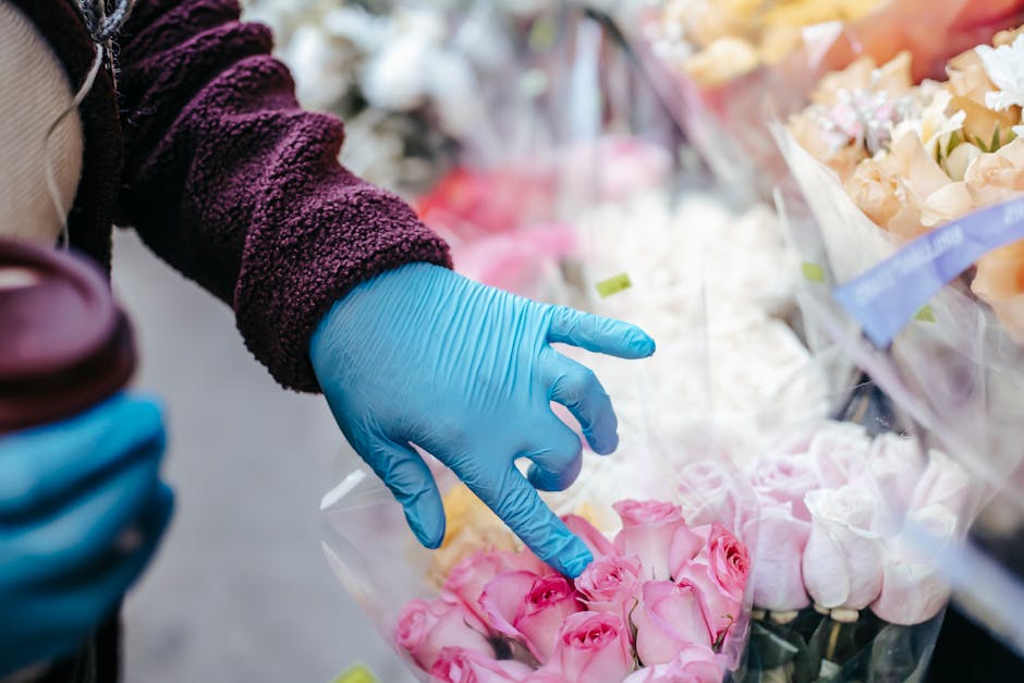 A person wearing a purple textured jacket and blue disposable gloves selecting a bouquet of light pink roses with delicate petals and green stems from a display of various floral arrangements wrapped 
