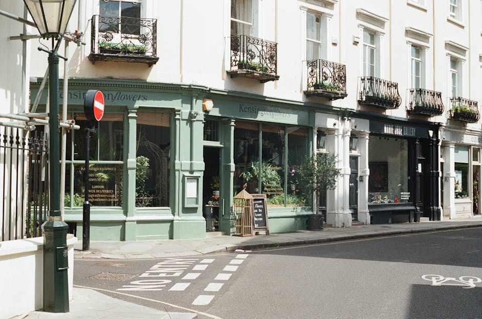 A storefront with large glass windows displaying various flowers and greenery inside, painted in a light green color with white accents. The shop has a black sign with white lettering reading 'Kensing