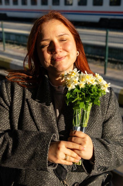 A woman with red hair, dressed in a gray coat, is holding a bouquet of fresh yellow and white daffodils with green stems and leaves, wrapped in clear plastic. She is outdoors, with sunlight illuminati