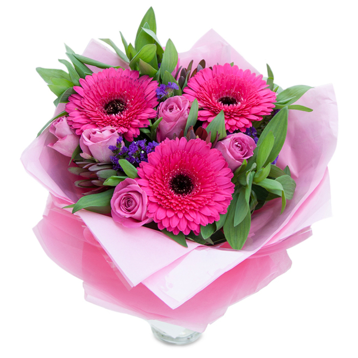 I picture this bouquet as a delicate balance of playfulness and grace, the kind of arrangement you might spot being carried along Brondesbury High Road on a sunny afternoon. Three vibrant fuchsia Gerbera daisies take centre stage, their many layered petals fanning out around deep dark centres that add dramatic contrast and visual rhythm. Interwoven close by are lavender-pink roses with petals that unfurl in soft, sculptural spirals-each one looking freshly cut, still holding that cool, dewy petal curl. Little clusters of purple statice tuck into the gaps, bringing pockets of tiny, papery blooms that offer both texture and a muted violet note to the palette. Rich green foliage-broad, glossy leaves that could be salal with the occasional eucalyptus stem-frames and lifts the bouquet, giving it natural body and a crisp, green scent.
The blooms are wrapped in an inner layer of pale pink tissue and finished with a translucent fuchsia-pink cellophane that crinkles pleasantly and catches light, suggesting the careful hands that folded it. The rim of a clear glass vase is visible at the bottom, so you see this piece as ready to be set on a hallway table in a Brondesbury home or to sit by a window overlooking Mapesbury Road. The overall effect is intimate and uplifting, with the roses offering soft perfume and the gerberas a subtle, fresh-green sweetness.
Made to celebrate moments both big and small-birthdays, anniversaries, or a spontaneous gift to cheer a friend-this bouquet brings a touch of neighbourhood warmth and floral craftsmanship to homes throughout Brondesbury and nearby Kilburn Park.